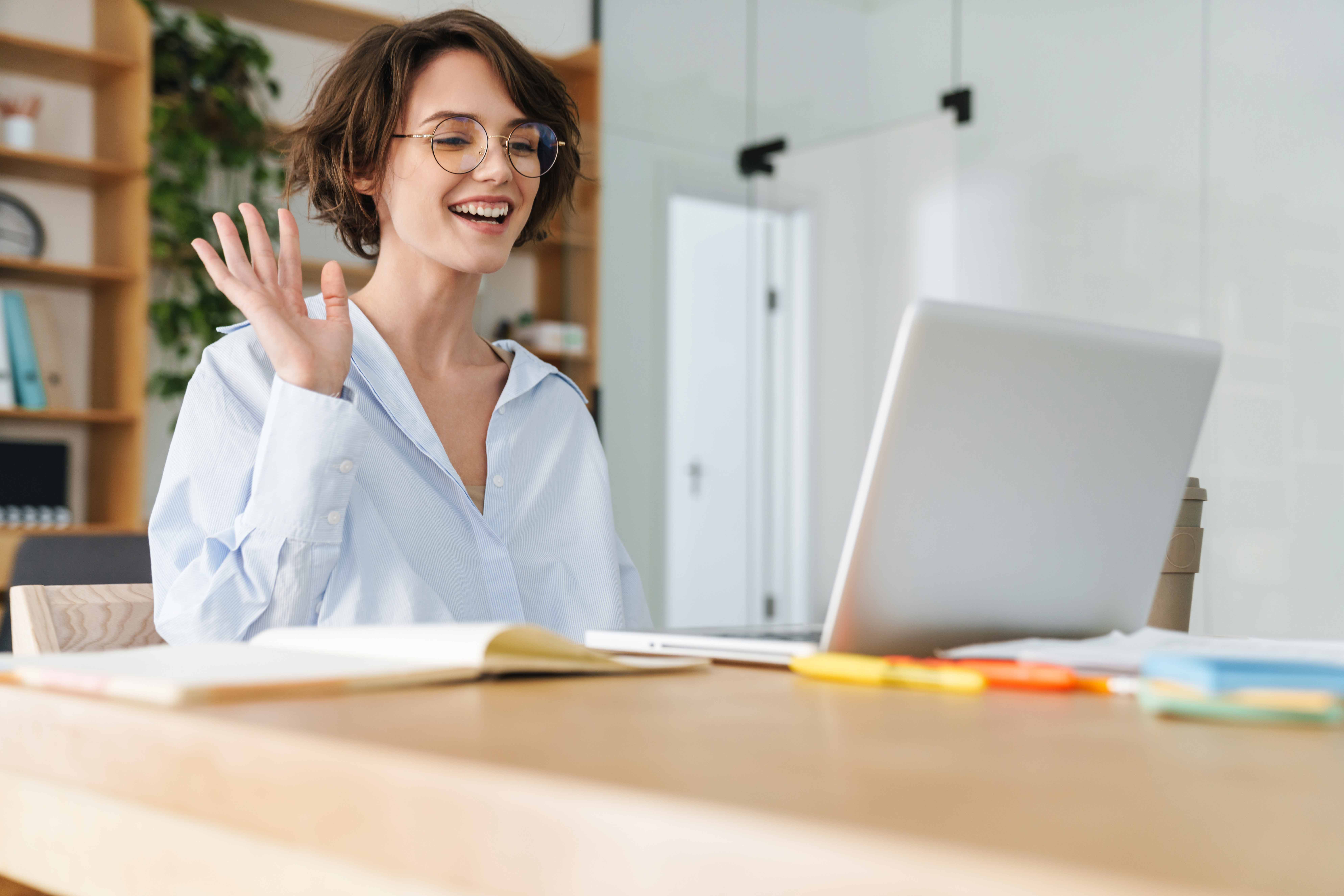 Das Bild zeigt eine junge Frau mit Brille, die vor ihrem Laptop sitzt. Ihre rechte Hand hebt sie etwas in die Höhe und lächelt dabei. Im Hintergrund sich unscharf Reagle und auch Blumen zu erkennen.