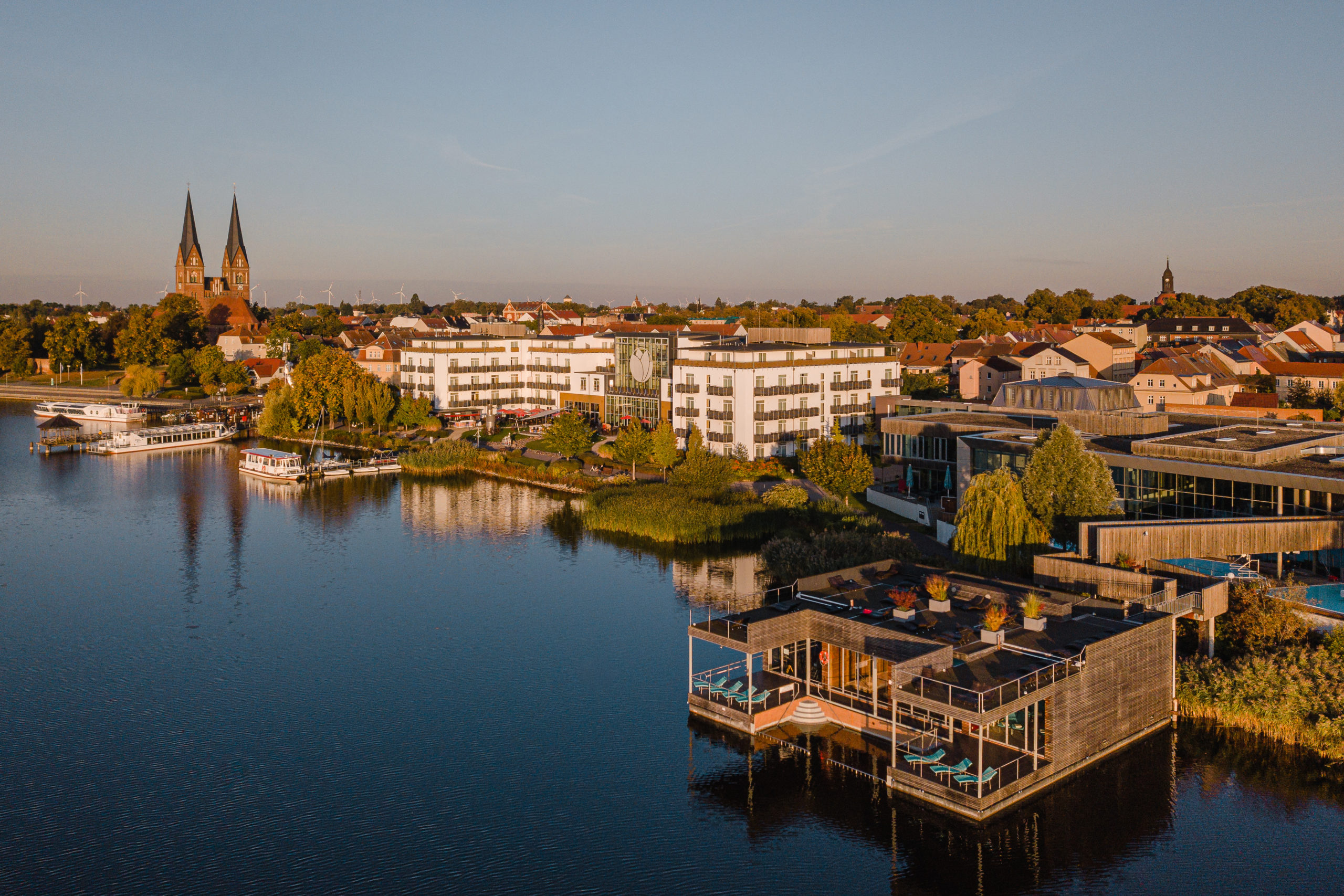 Das Bild zeigt in einer Luftaufnahme die Uferseite von Neuruppin im Sonnenaufgang. rechts beginnt die Pfarrkirche, weiter entlang des Ufers sieht man das Resort Mark Brandenburg und die Therme. Im Anschnitt sieht man unten den Ruppiner See.