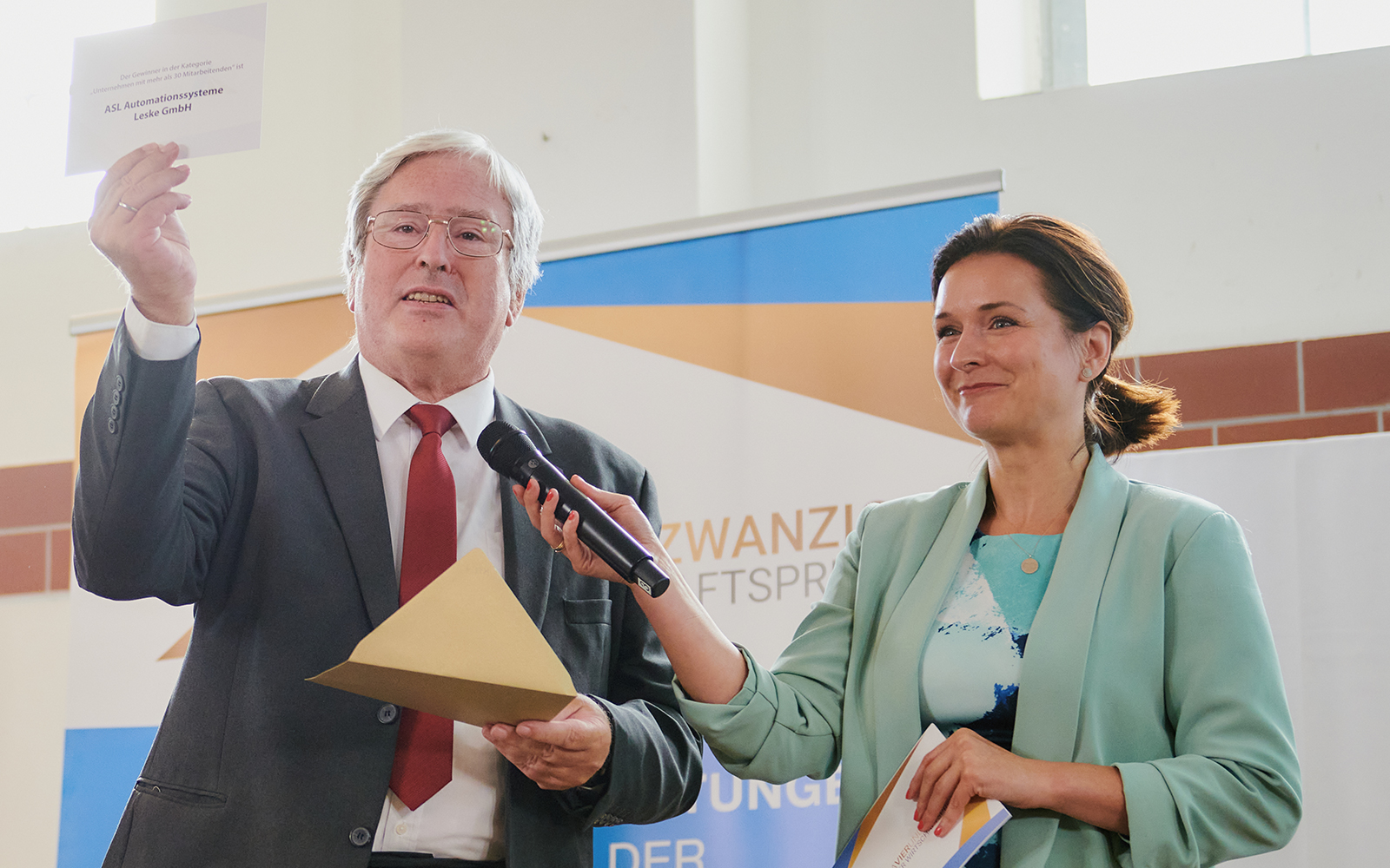 Das Foto zeigt den ehemaligen Wirtschaftsminister Jörg Stenbach bei der Bekanntgabe der Gewinner des AVIERUNDZWANZG Wirtschaftspreies 2022. Moderatorin Tina Knop hält ihm des Mikrofon hin, während er den Zettel mit dem Gewinner ASL Automationssysteme Leske GmbH in die Höhe hält.