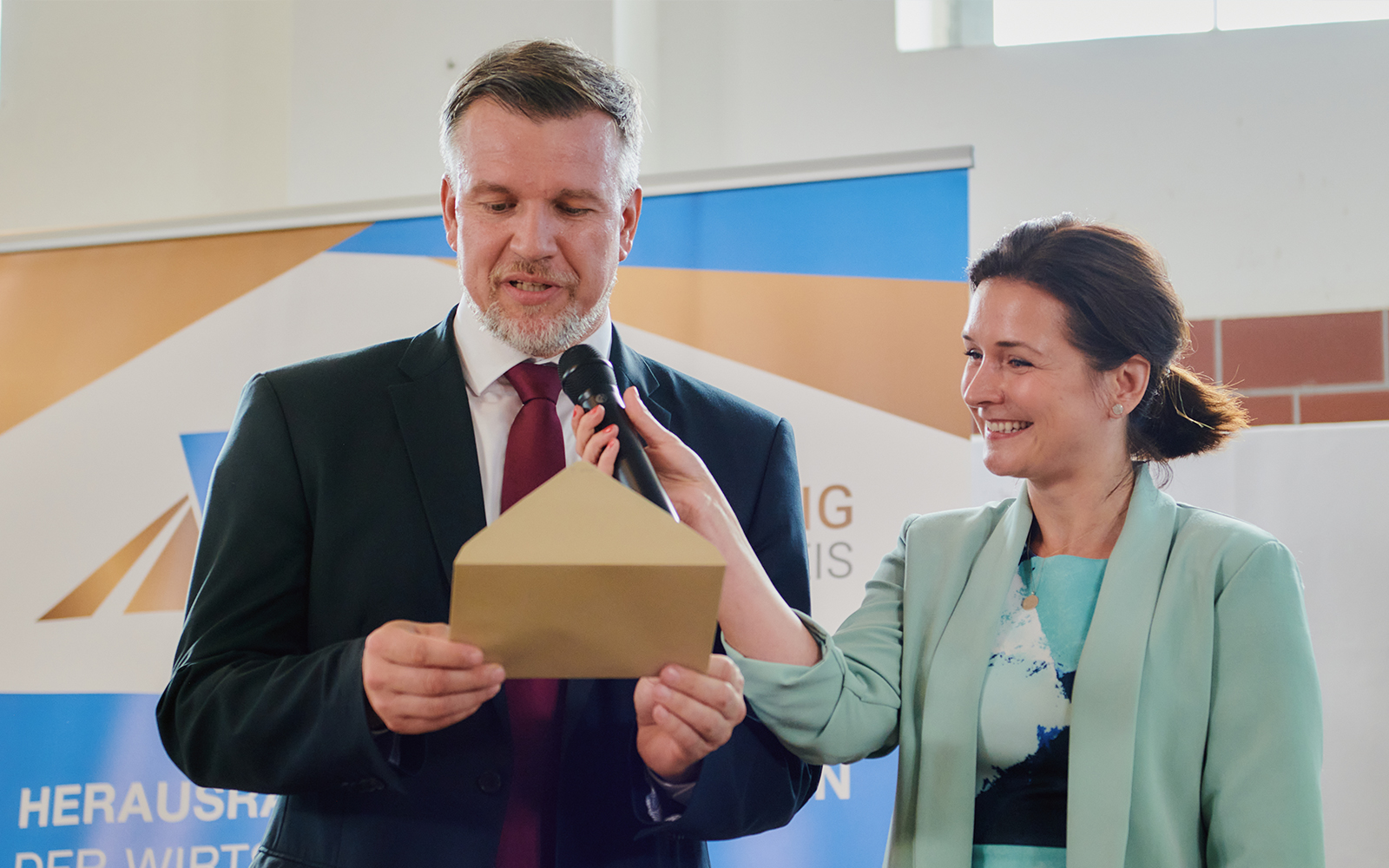 Das Foto zeigt Landrat Ralf Reinhardt. Landrat des Landkreies Ostprignitz-Ruppin bei der Bekanntgabe der Gewinner des AVIERUNDZWANZG Wirtschaftspreies 2022. Moderatorin Tina Knop hält ihm des Mikrofon hin, während er den Namen eines Gewinners aus einem goldenen Umschlag vorließt.