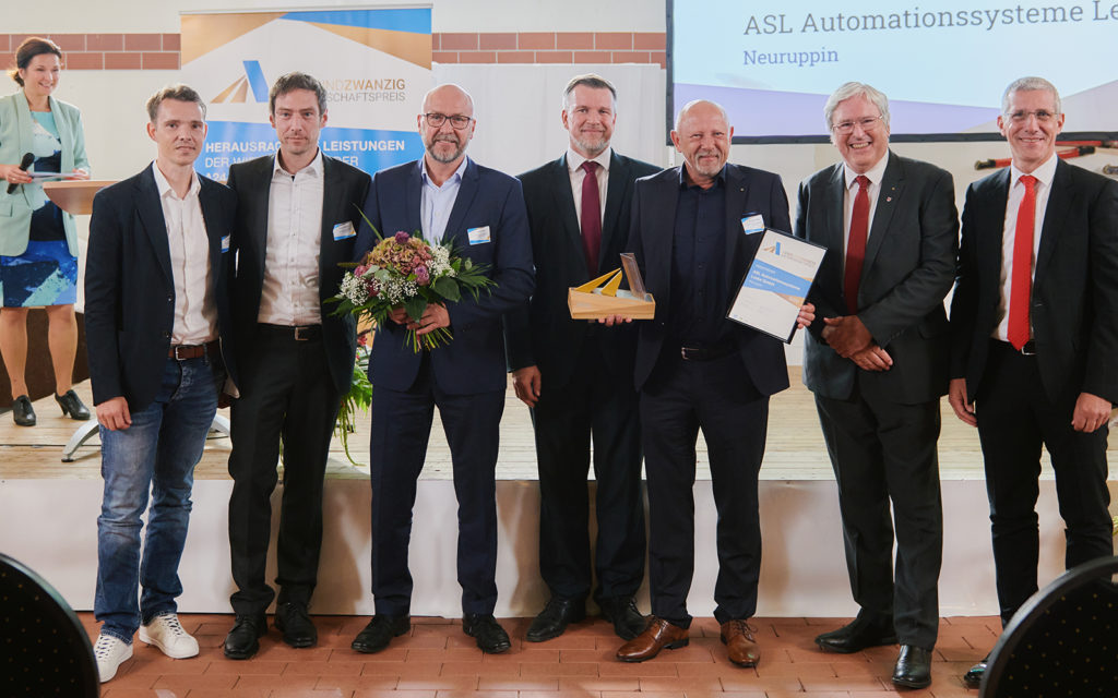 Das Foto zeigt sieben Menschen, die nach der Preisverleihung des AVIERUNDZWANZIG Wirtschaftspreises 2022 in die Kamera schauen. Die drei linken Personen sind von der ASL Automationssysteme Leske GmbH. In der Mitte steht Ralf Reinhardt (Landrat des Landkreises Ostprignitz-Ruppin). Der dritte von rechts ist Andreas Leske (Geschäftsführer der ASL Automationssysteme Leske GmbH), er hält den Preis und die Siegerurkunde in den Händen. Der zweite von rechts ist der damaligen Wirtschaftsminister Jörg Steinbach und ganz rechts steht Markus Rück (Vorstandsvorsitzender der Sparkasse Ostprignitz-Ruppin).