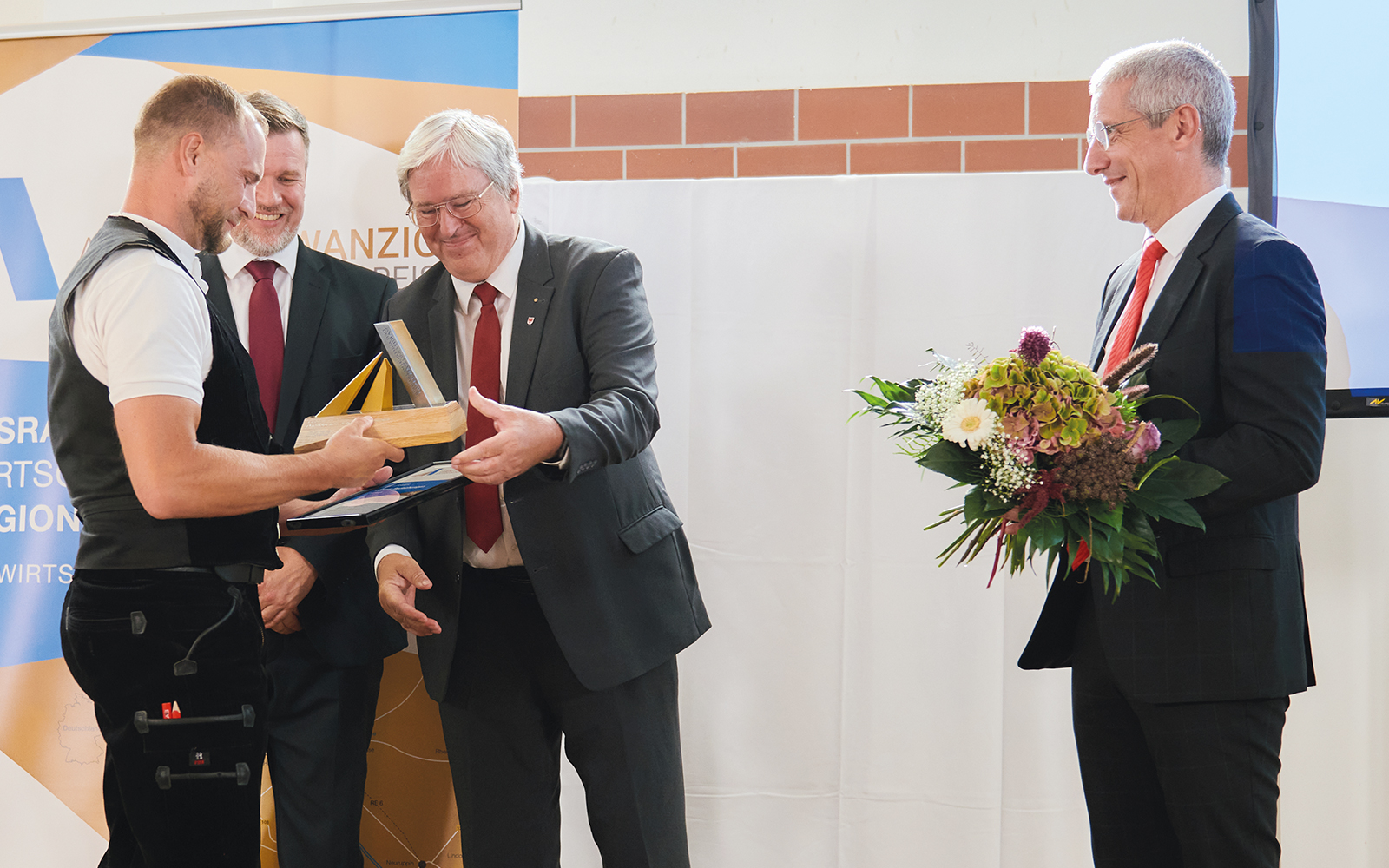 Das Foto zeigt die Preisübergabe an den Preisträger des AVIERUNDZWANZIG Sonderpreises 2022 Marcel Lublow (links). Der ehemalige Brandenburger Wirtschaftsminister Jörg Steinbach (Mitte) überreicht ihm die Preisfigur auf der Bühne. Im rechten Bildrand steht Markus Rück (Vorstandsvorsitzender der Sparkasse Ostprignitz-Ruppin) mit Blumen für den Preisträger.